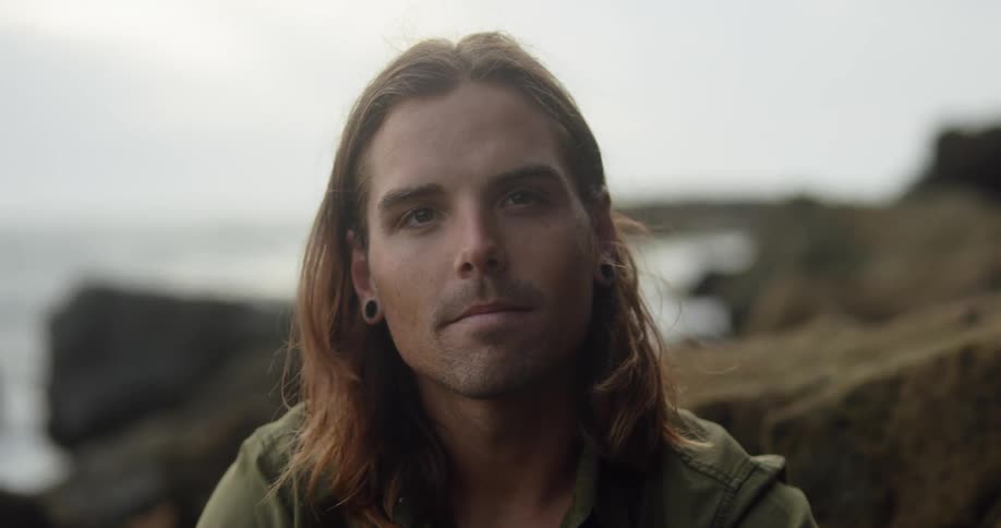 Long haired young man sitting on the rocks on the beach looking at the camera at sunset