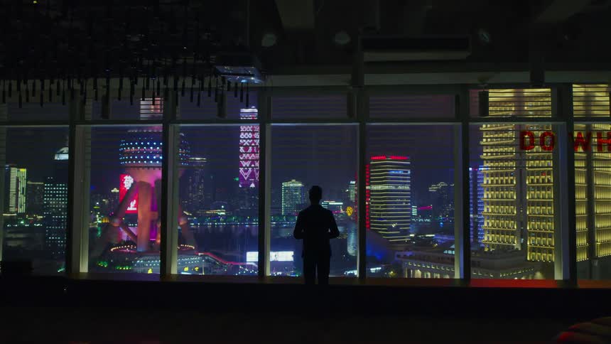 A man stands inside a building and looks outside large windows at the night skyline