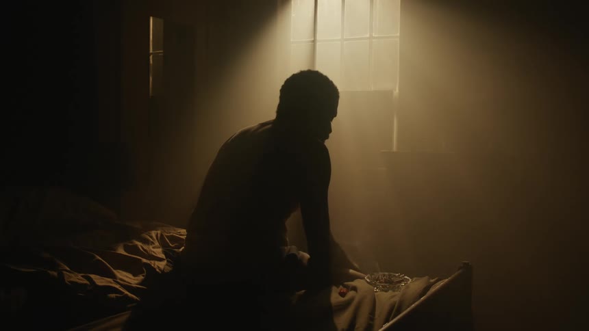 Man silhouetted by light from window sits on bed with hunched shoulders as he light then smokes a cigarette