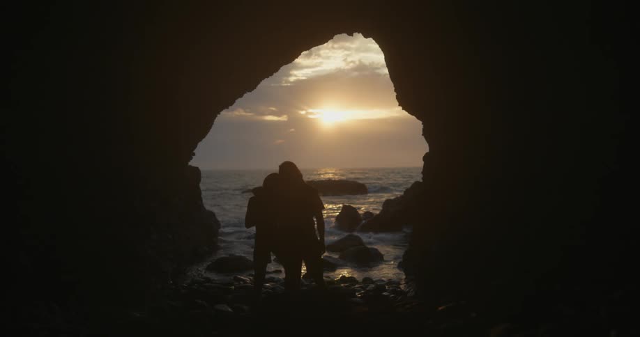 Couple stand with arms around each other staring at horizon and sun as ocean waves crash against rocks at mouth of sea cave