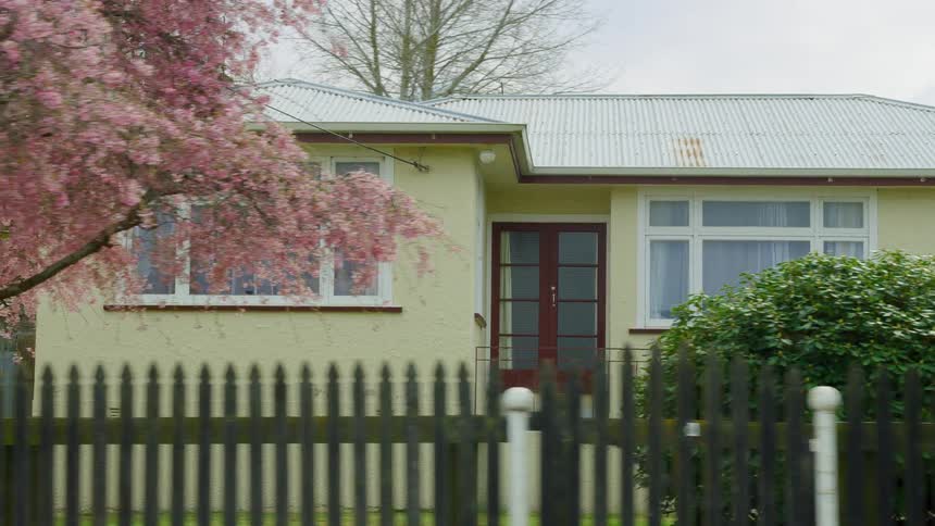A fence surrounds a flowering tree and bush in the front yard of a yellow house