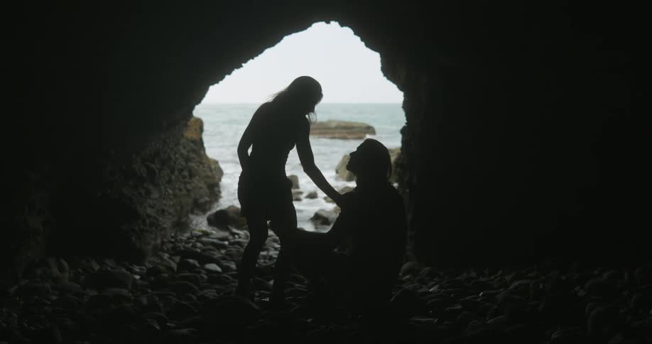 A woman looks at the man sitting on the rocks then sits beside him holding his arm as waves splash at the cave opening