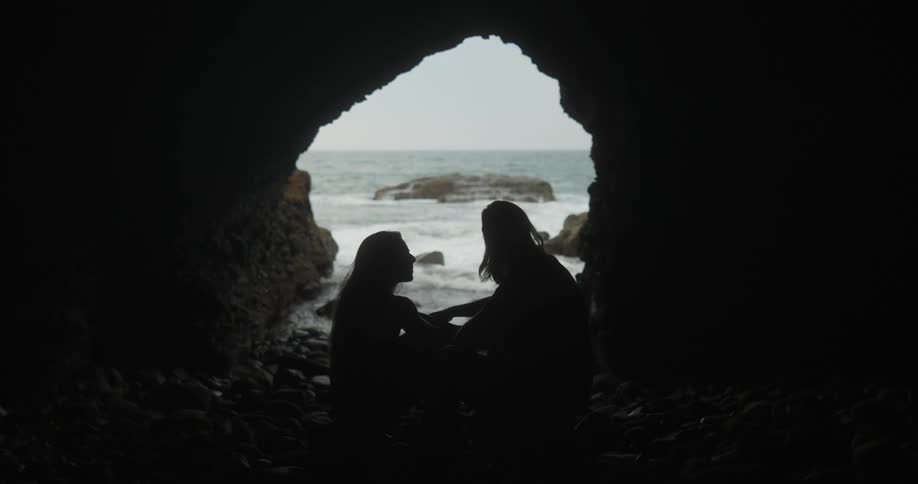 A silhouette of a man and woman in a cave with the ocean sky and clouds in the background