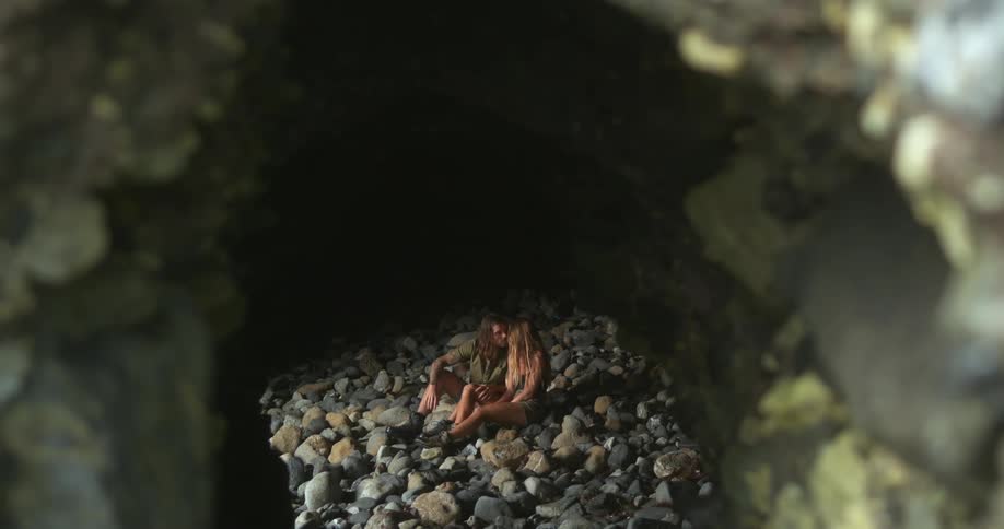 Man with long hair and woman with brown hair sits on stones in dark cave talking to each other
