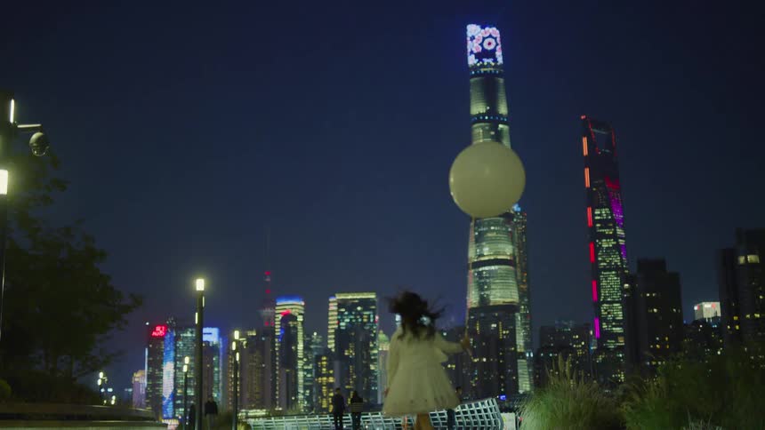 A young girl holding a balloon skips down a walkway in a metropolis at night