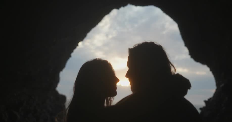 A man and woman stand on the shore and hug and kiss as the sun sets