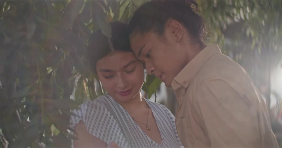 Sun shines through tree leaves as couple looks into each others faces and lean close to touch foreheads