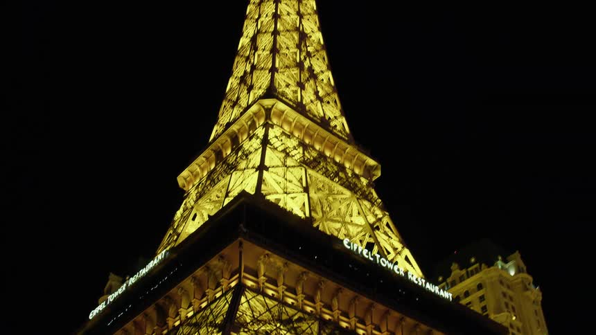 Statues sit at base of eiffel tower restaurant bordered by other buildings in city at night