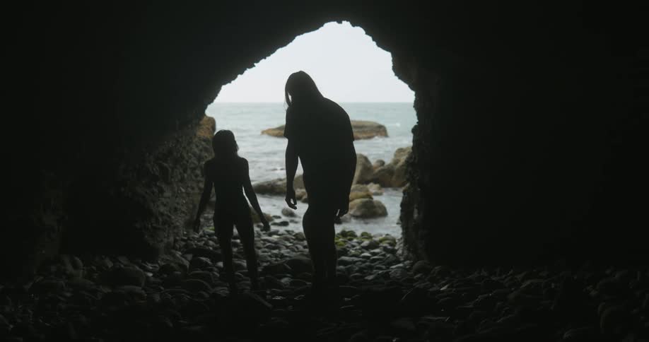 A man and woman walk through the entrance of a cave along the coast and sit on the rocks