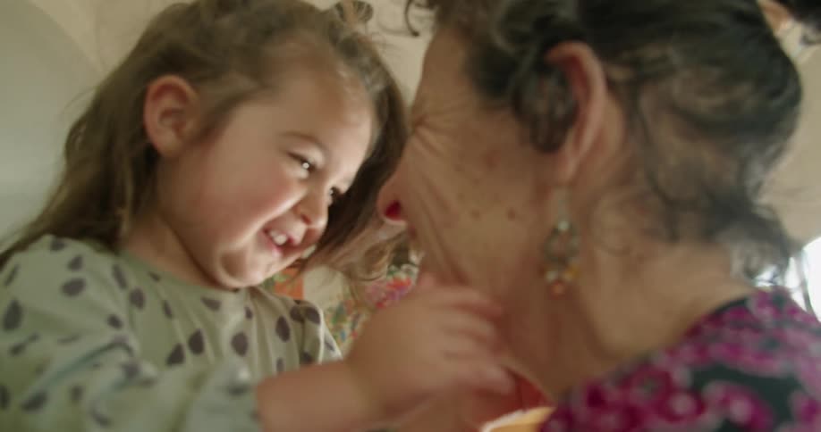 A little girl playing with a womans face in a room with a wall and window in the background