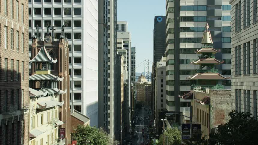 Traffic moves along downtown city streets near buildings with pagoda architecture and church with suspension bridge in the distance