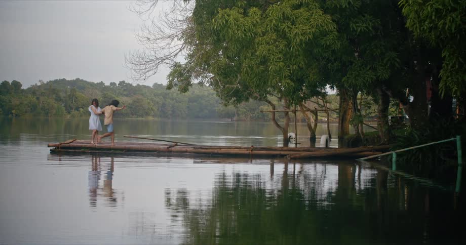A man runs around a woman holding onto his arm on a pier made of logs on a lake on a cloudy day