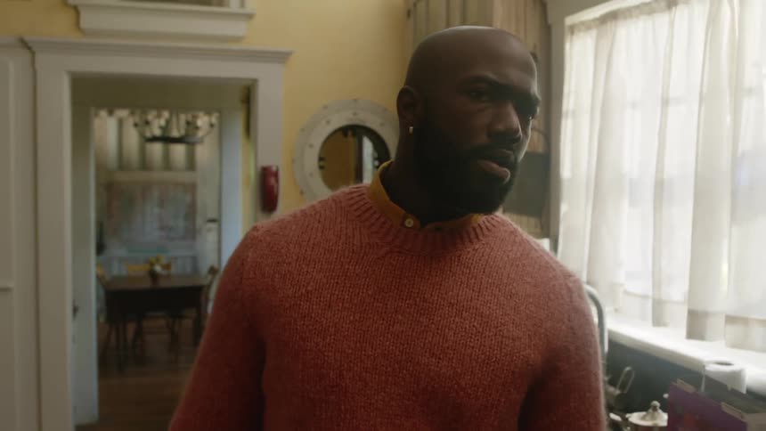 A man with a sweater in a kitchen with a window a sink and a faucet in the background