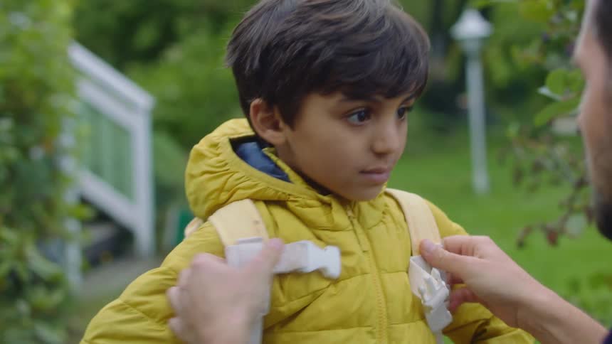 A man putting on a jacket and a backpack on a boy with a house in the background