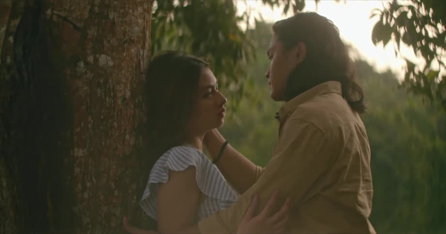 A man hugging and caressing a woman underneath a tree with the sky and clouds in the background