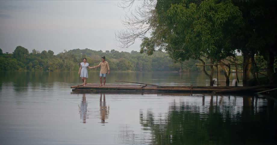 Man holds womans hand to help her maintain balance as they walk to edge of pier on rippling water reflecting them and the nearby trees under a cloudy sky