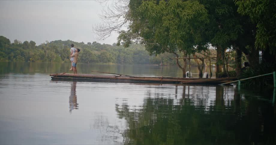 A man lifts a woman into the air while standing on pier made of logs on a lake during the day