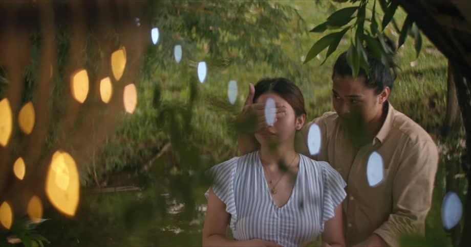 Man covers womans eyes escorting her to decorated trees on edge of stream before letting her look