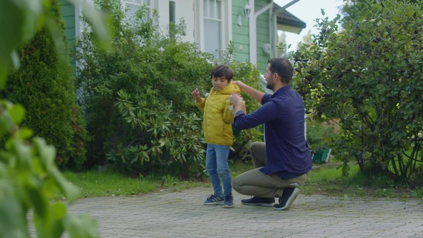 A man putting on a jacket and a backpack on a boy with a house the sky and clouds in the background