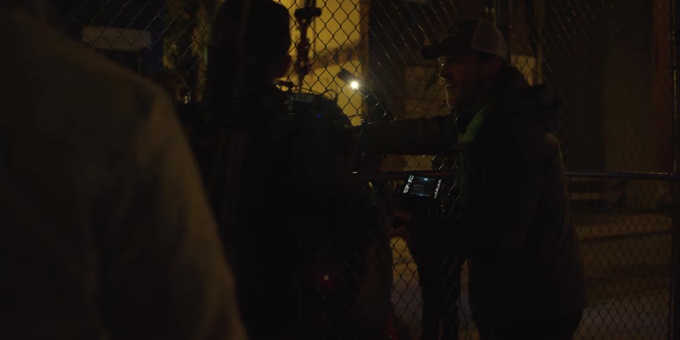 A cameraman and photographers film a chaotic scene near a chain link fence at night