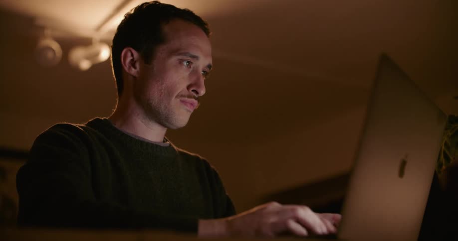 A man in a room using a laptop with a wall and picture frame in the background
