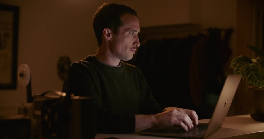A man types on his laptop in a dimly lit room at home