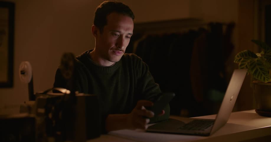 A man is sitting at a desk scrolling on a cell phone and typing on a laptop
