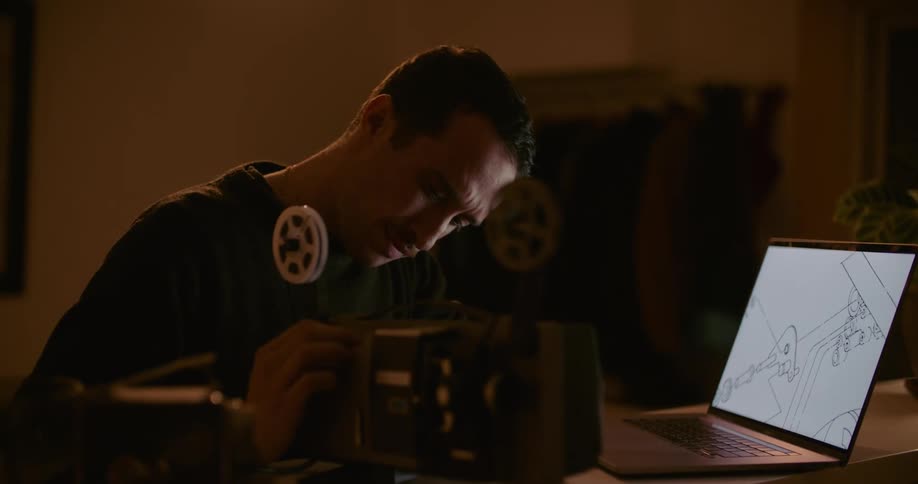 A man in a room using a laptop and adjusting a projector with a wall in the background