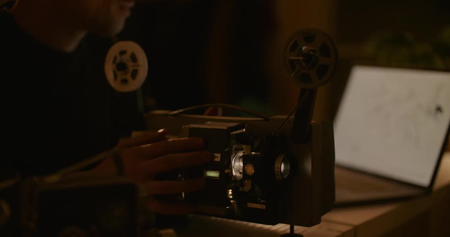 Man sits at table working with film projector as he consults diagram on laptop screen