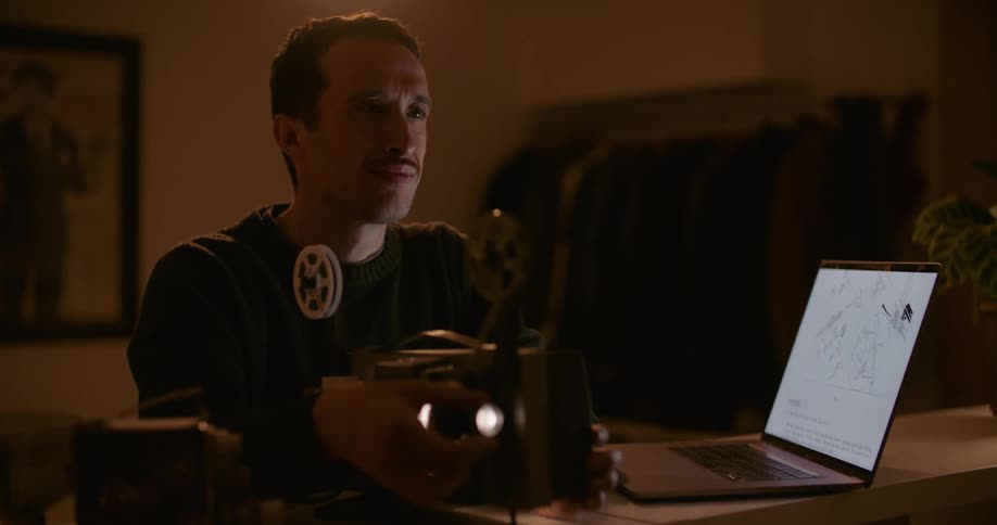 A man in a room looking at a laptop and adjusting a projector with a wall in the background