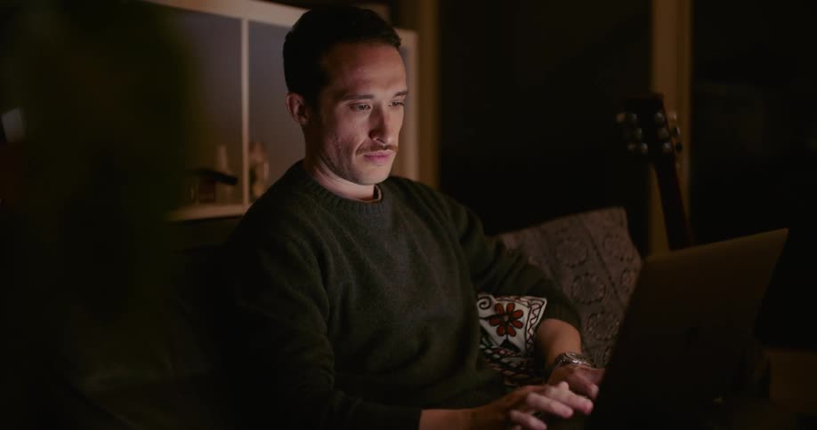 A man in a room sitting on a couch typing on a laptop with a guitar and a wall in the background