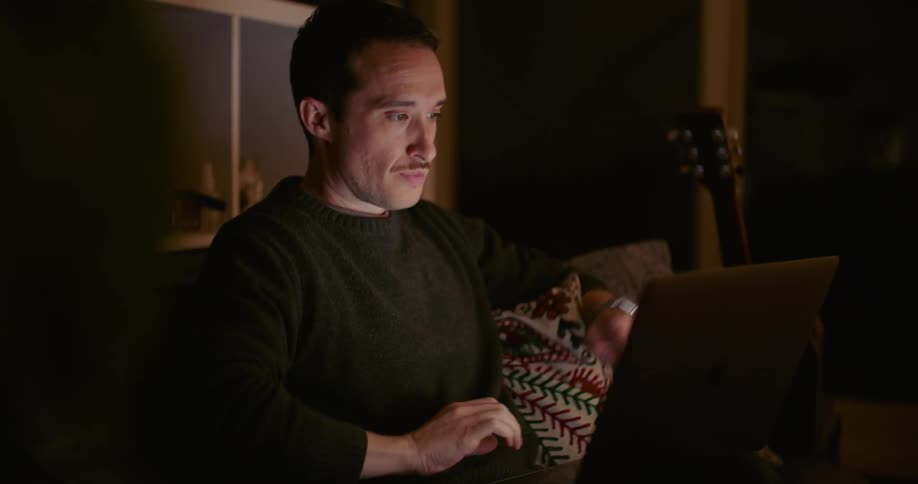 Man with mustache and black sweater uses smartphone and types on laptop in dark room