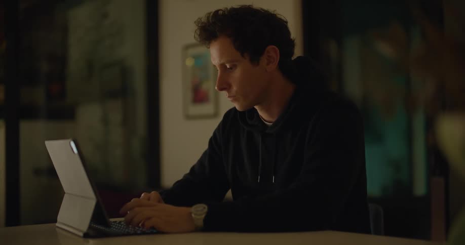 Young man sitting at a desktop typing on a laptop inside a dimly lit room