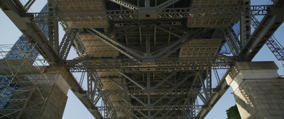 Under a clear blue sky stands an enormous metal bridge as seen from below