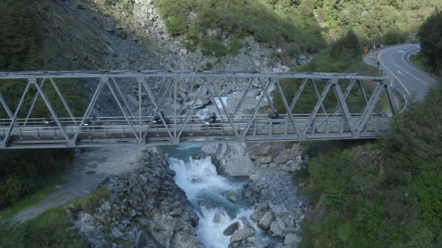 A group of motorcyclist drive across a bridge spanning a ravine