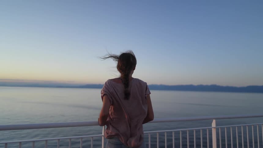 A woman stands against a railing and watches the ocean as wind blows through her hair