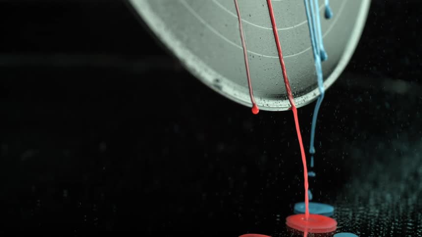 Red and blue paint drip down metal disk falling onto dark surface along with floating particles