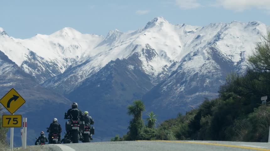 Group of motorcyclists ride along road past scrub covered slope with snow covered mountains on horizon