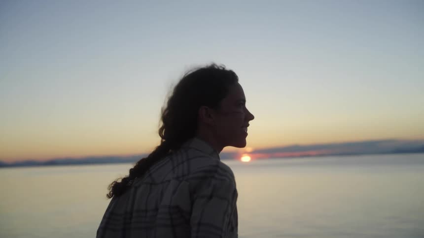 A woman smiles to herself as wind blows through her hair as she watches the sun set over the sea