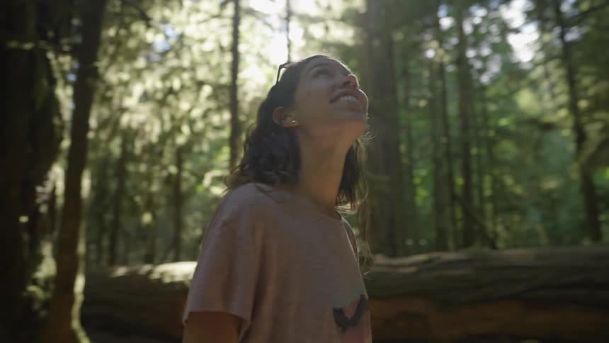 A smiling young woman looks around at her surroundings while walking through a forest during the day