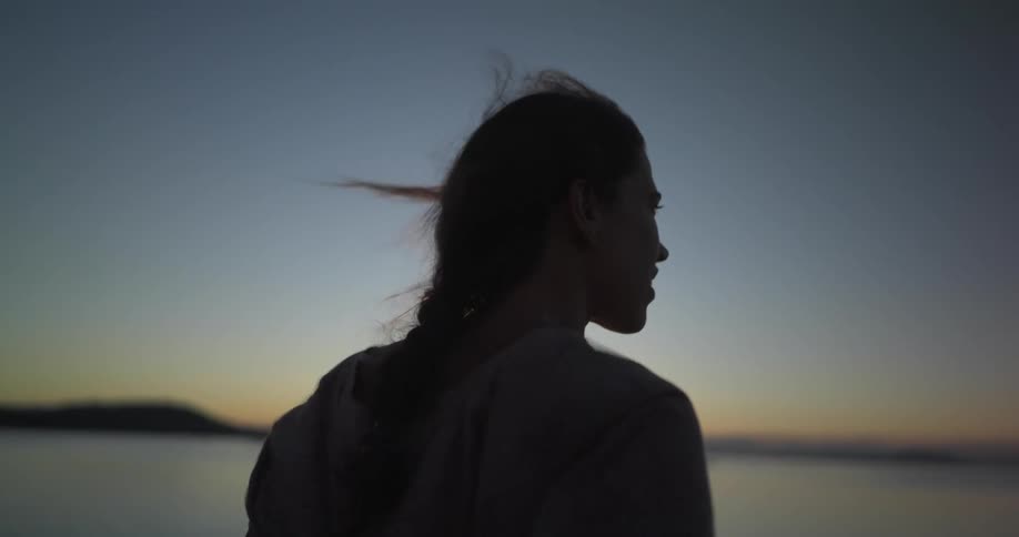 A silhouette of a woman with wind blowing in her hair with a lake the sky and clouds in the background