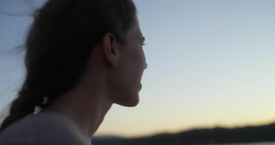 Woman stands on shore on windy day smiling as she looks out toward red tinted horizon