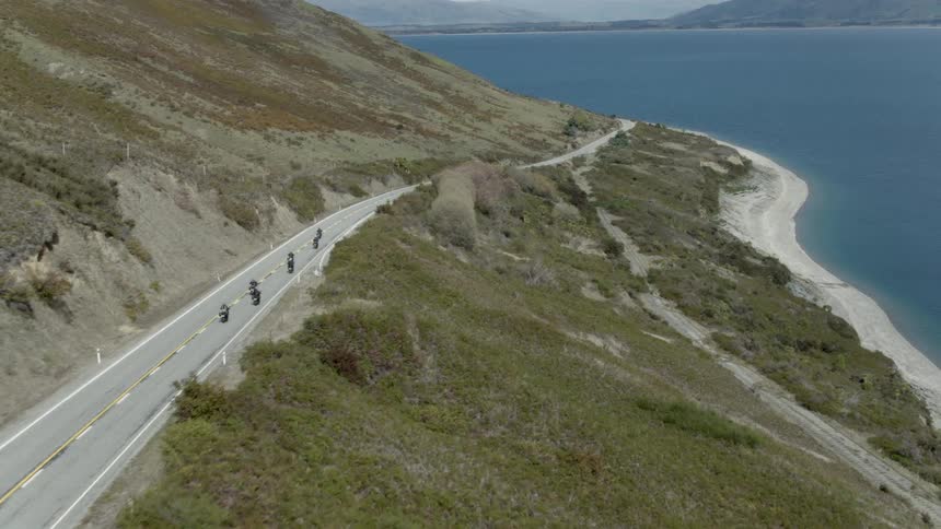A line of motorcycles travels on a coastal highway with slopes leading to sandy beaches