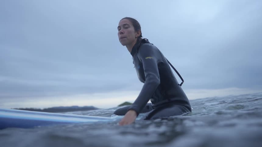 Woman wearing wet suit sits up on surfboard and starts paddling as she looks around