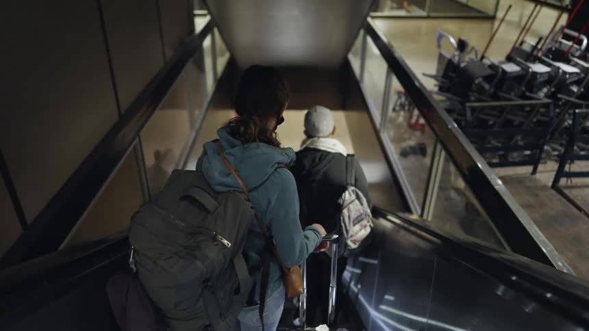 Two people ride down an escalator in an airport holding onto luggage