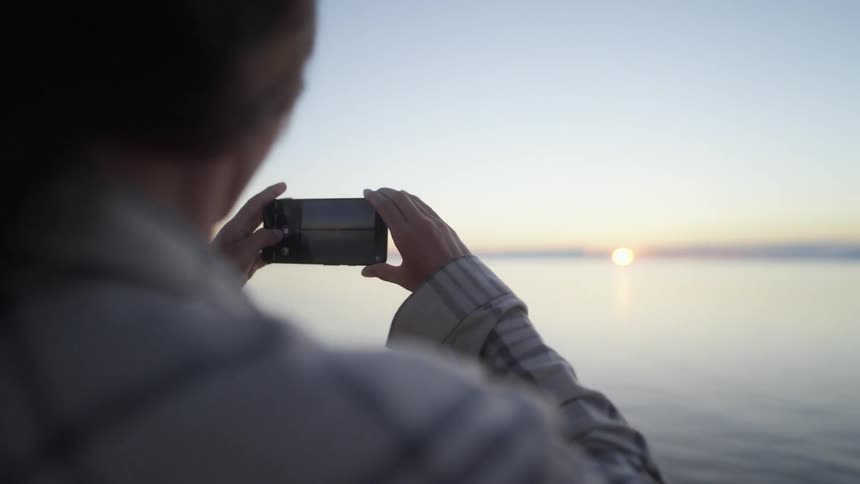 Woman with brown hair and shirt takes photograph of the sea and sunset while traveling by ferry boat
