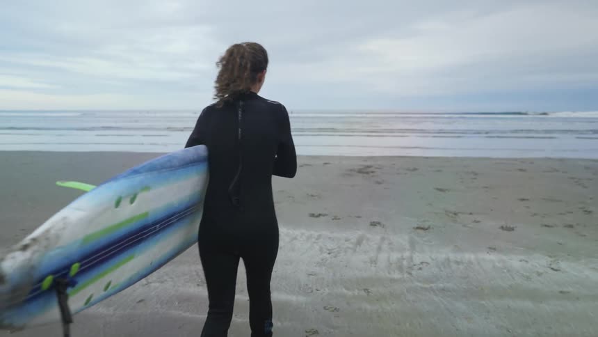 A woman with a wetsuit holding a surfboard and walking on a beach with the ocean sky and clouds in the background