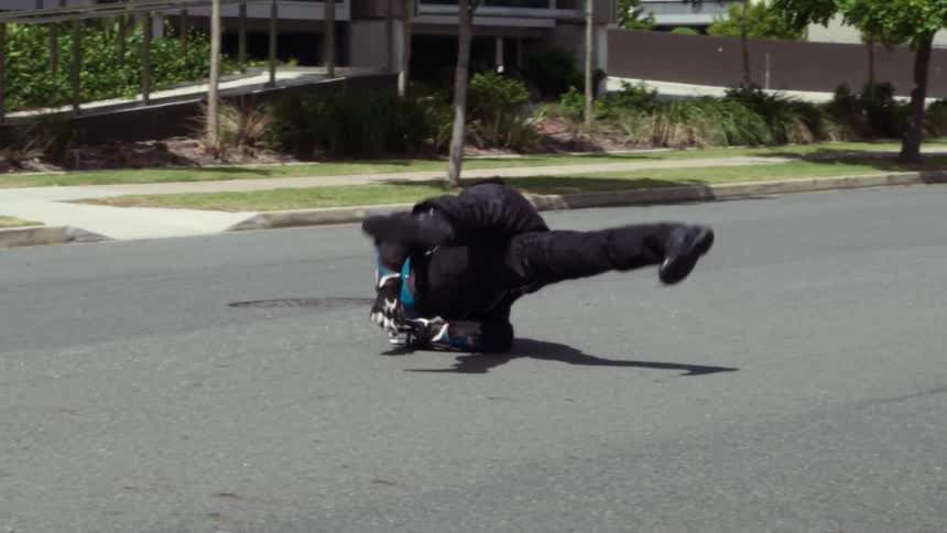 A man in a helmet and protective riding clothing spins and rolls in pain down a neighborhood street