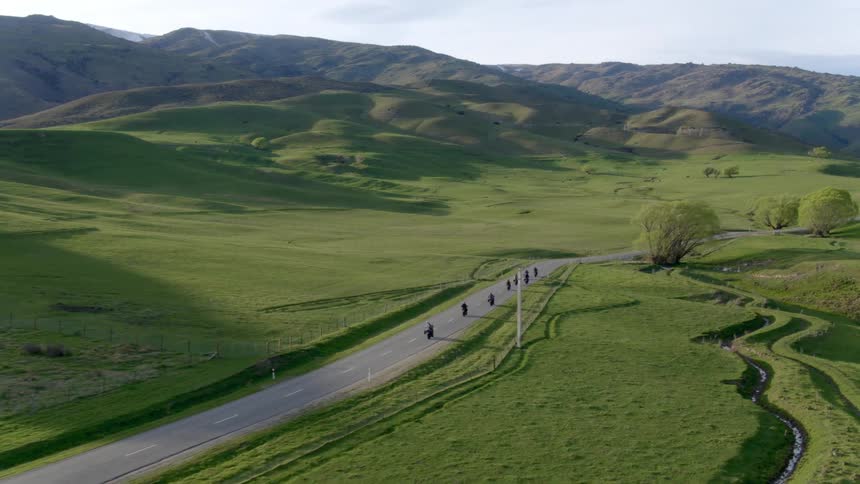 A group of motorcyclists travel in a line along a road through the countryside on a bright day