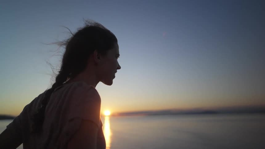 Smiling woman stands hair blowing in wind looking over ocean as water reflects sun sitting on horizon